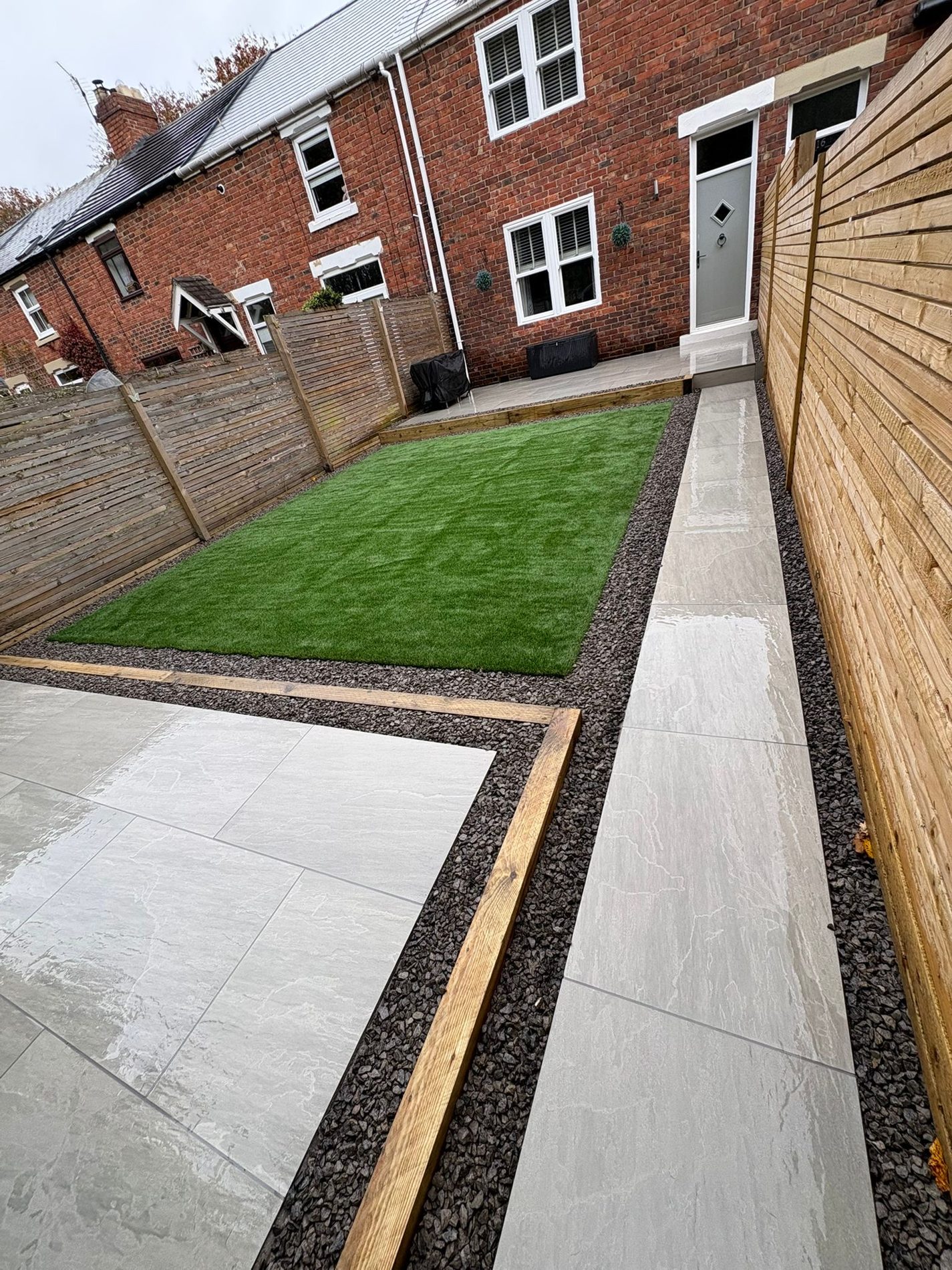 A narrow terraced garden with a porcelain path, timber fencing and an artificial lawn