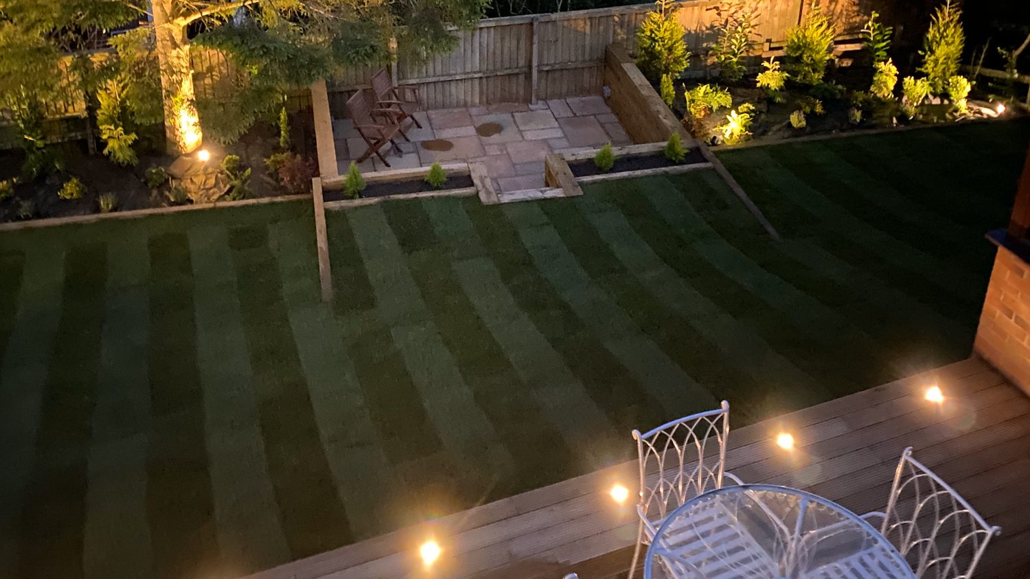 A garden at dusk with a striped lawn, deck lights and a seating area